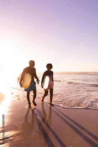 Happy african american couple with surfboards walking at shore against clear sky at sunset