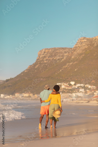 Rear view of african american couple with arms around walking at beach against mountain and blue sky