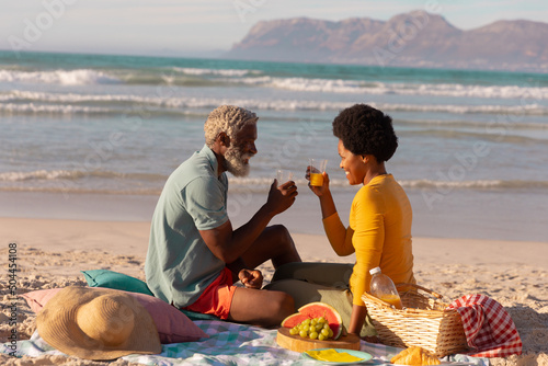African american couple toasting juice while sitting with food on blanket against sea at sunset
