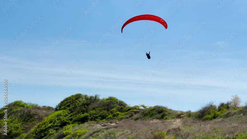 A paraglider soars above the sea beach on a summer sunny day. The red wing of the parachute and the pilot on the background of the sea coast with a sandy beach and blue sky. A paraglider soars above the sea beach on a summer sunny day. The red wing of the parachute and the pilot on the background of the sea coast with a sandy beach and blue sky.