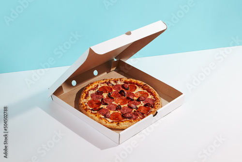 Italian pizza in paper box on coloured background. Meat pizza with sausage and salami in delivery box in minimal style . American pizza delivery concept with color backdrop.