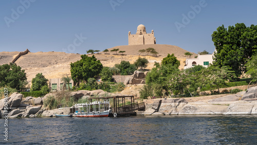 There are picturesque boulders on the river bank, a tourist boat is moored. At the top of a sand dune, against the blue sky, stands the ancient mausoleum of the Aga Khan. Egypt. Nile. Aswan photo