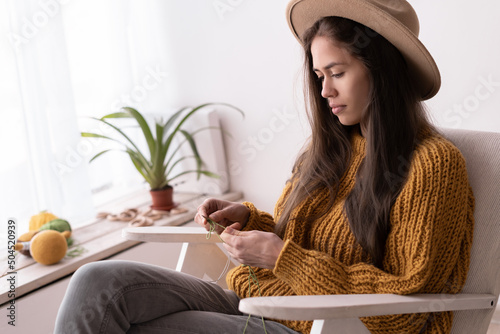 young female knitter in knitted warm sweater enjoys knitting on armchair at home in cozy winter time, knits crochet, Home comfort, hobby and needlework photo