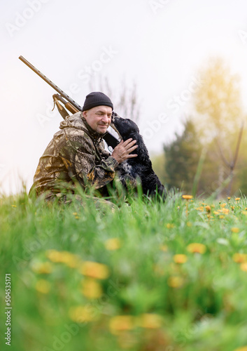 Male hunter with a gun on his shoulder hugging a Russian spaniel dog at sunrise in the woods