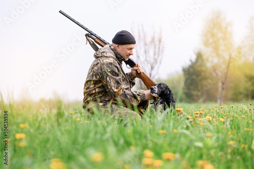 Male hunter with a gun on his shoulder stroking a dog Russian spaniel at sunrise in the woods