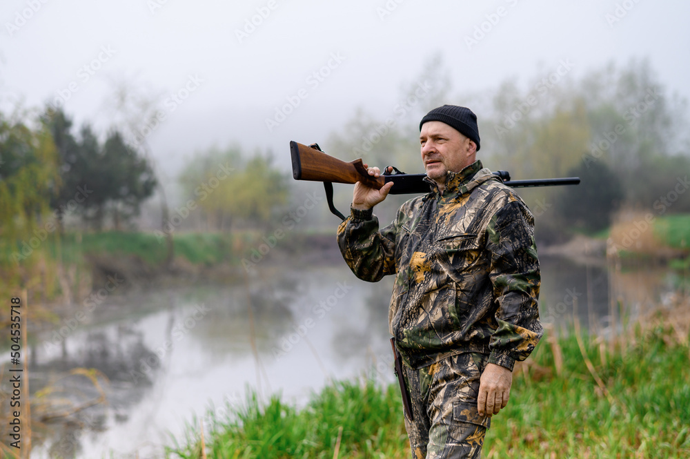 Fototapeta premium A handsome man with a gun on his shoulder looks at the lake in the fog. Medium plan