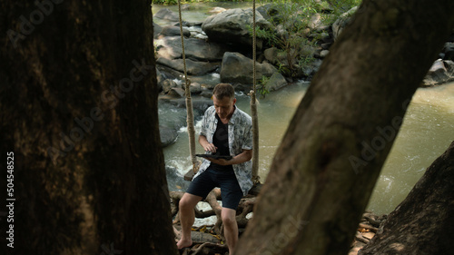 A middle-aged man in the middle of the forest, trees, nature and using a laptop.