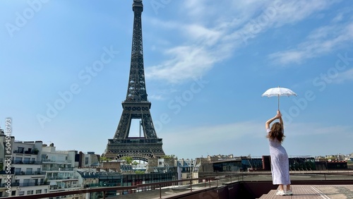 a beautiful young girl in a summer dress stands against the backdrop of the Eiffel Tower with a lace umbrella, she raised her hand and as if taking off there is a place for advertising travel agency.