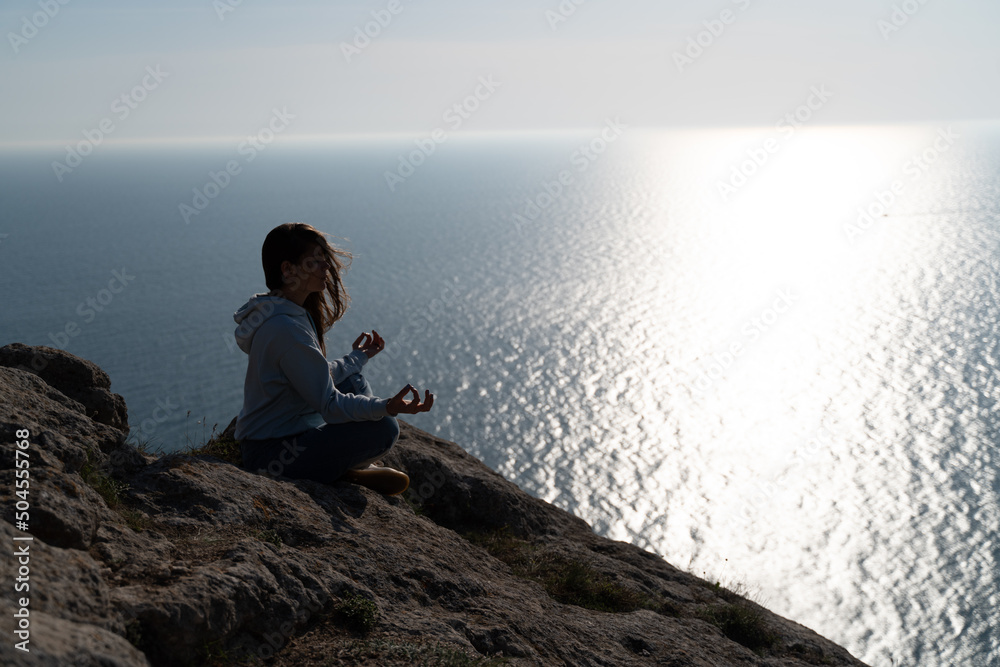 Woman tourist enjoying the sunset over the sea mountain landscape. Sits outdoors on a rock above the sea. She is wearing jeans and a blue hoodie. Healthy lifestyle, harmony and meditation