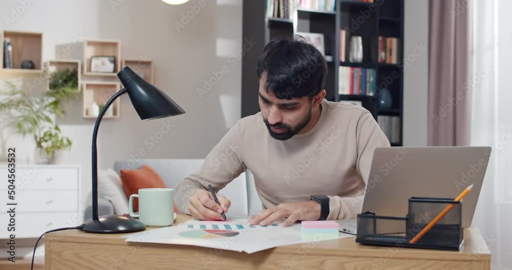Arabian young man sitting at desk and working with diagram, charts and graphics at laptop computer. Arab male checking numbers and figures in business at home. Freelancing job. Phone talk. Arabian young man sitting at desk and working with diagram, charts and graphics at laptop computer. Arab male checking numbers and figures in business at home. Freelancing job. Phone talk.