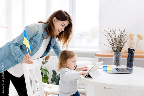 Young smiling mom works at home with her cute baby. Work at home, the girl communicates with her family at a distance from Ukraine. Peace of Ukraine. Blue Yellow. Patriot of his country.