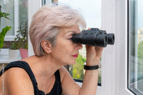 Portrait of an elderly woman looking through binoculars at other people's house windows and the street. Concept: spying on neighbors, gathering information, espionage and gossip.