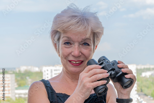 Portrait of an elderly, fastidious woman of 60-65 years with binoculars against the backdrop of the urban landscape.Concept: spying on neighbors, gathering information, espionage and gossip. photo