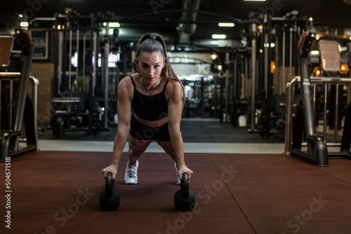 Athletic woman using kettlebells while doing plank in gym