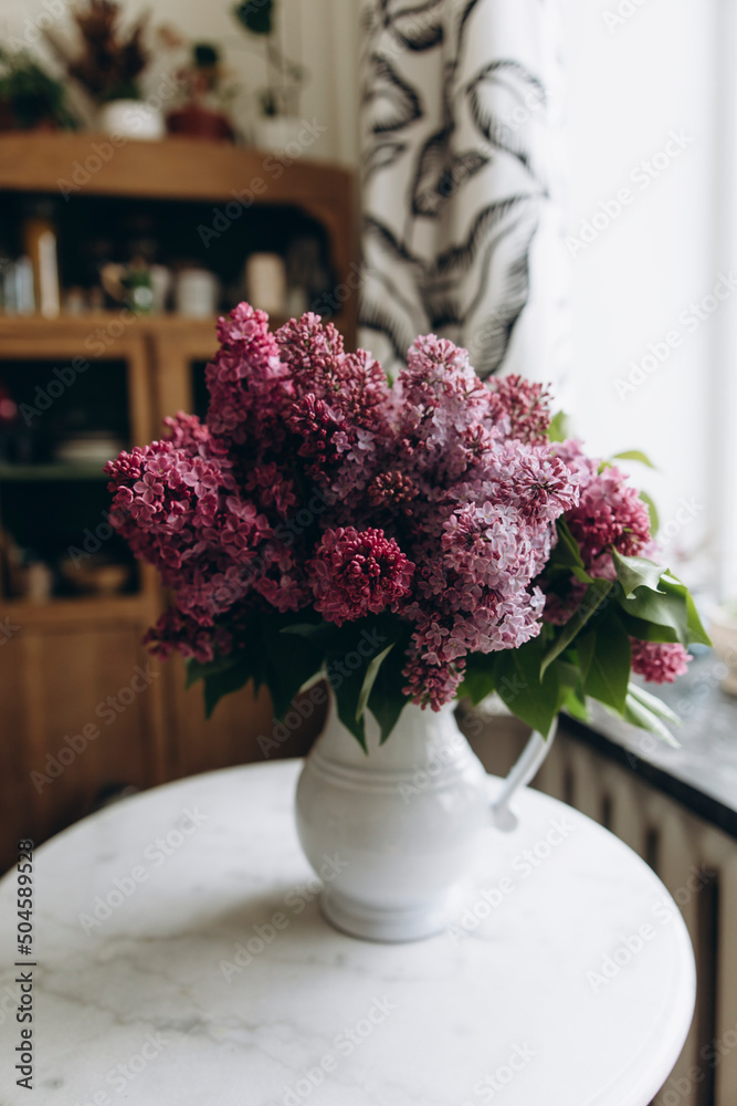 a bouquet of lilacs in a white vase on the table. lilac branches in a ceramic vase on a white table in the kitchen. a fresh bouquet of lilac decorates the kitchen room a bouquet of lilacs in a white vase on the table. lilac branches in a ceramic vase on a white table in the kitchen. a fresh bouquet of lilac decorates the kitchen room