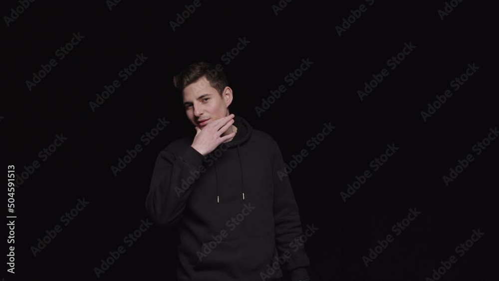 A young guy in a black hoodie on a black background with a brazen grin rubs his chin. A young guy in a black hoodie on a black background with a brazen grin rubs his chin.