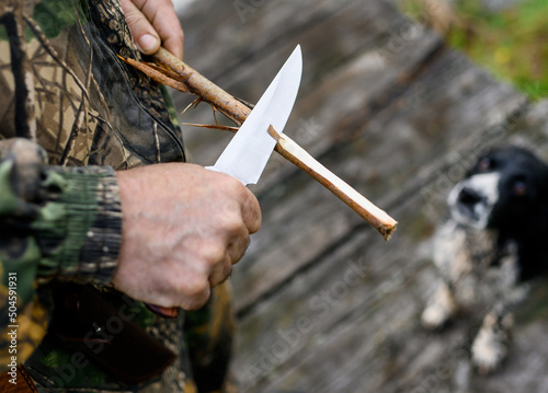 Hunter planes a branch with a knife on the background of a dog and a bridge. High quality photo