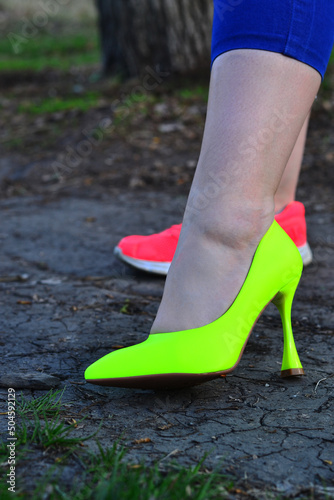 Legs of a girl in different shoes. Bright shoes with heels and sports shoes. Against the background of asphalt and grass.