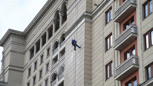 Male industrial climber in blue uniform cleans a wall of building using a high-pressure water spray. Real time video. High rise cleaning theme.