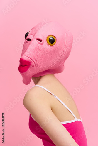 Skinny woman in a pink Halloween costume with a fish head on her face poses funny against a pink background and looks at the camera  art concept photo