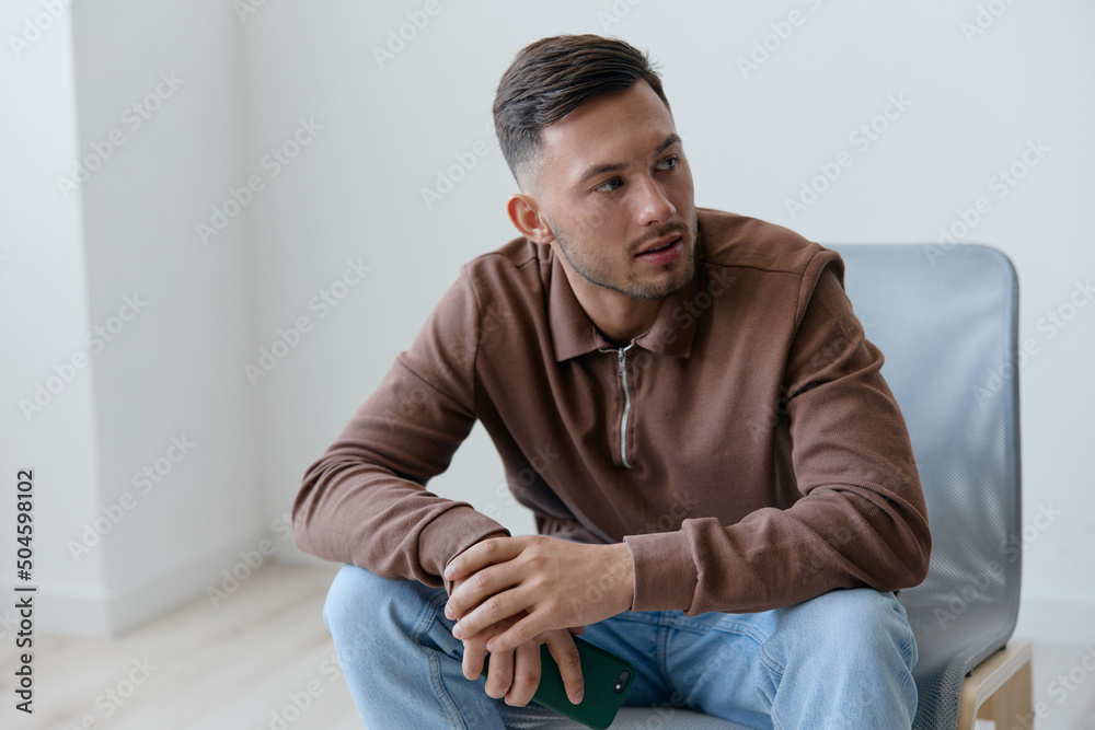 Happy young man holds phone looks aside sitting at chair in office on over white wall home background. Social Media concept. Copy space for ad. Mobile App offer Banner Mockup