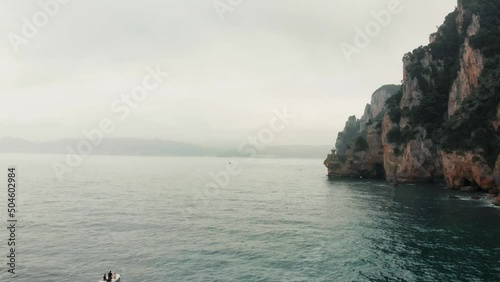 Small boat or yacht sailing alone on the sea near the cliffs where a lighthouse is located. Shot made with an aerial drone. Adventure of a cloudy day with waves.