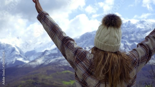 Stylish young girl wears a plaid shirt and a white fleece hat as she contemplates the views of the picos de europa natural park, she raises her arms and hands in euphoria. snowy mountains in winter.