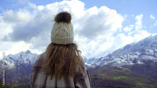 Stylish young girl wears a plaid shirt and a white fleece hat while looking at the views of the picos de europa natural park, some snowy mountains in winter on a sunny day with clouds.