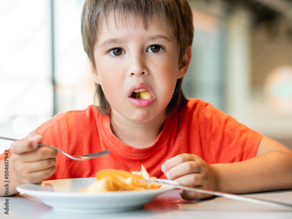 Kid in red t-shirt eats food with knife and fork. Fastidious boy with puzzled expression on face. Kid in red t-shirt eats food with knife and fork. Fastidious boy with puzzled expression on face.