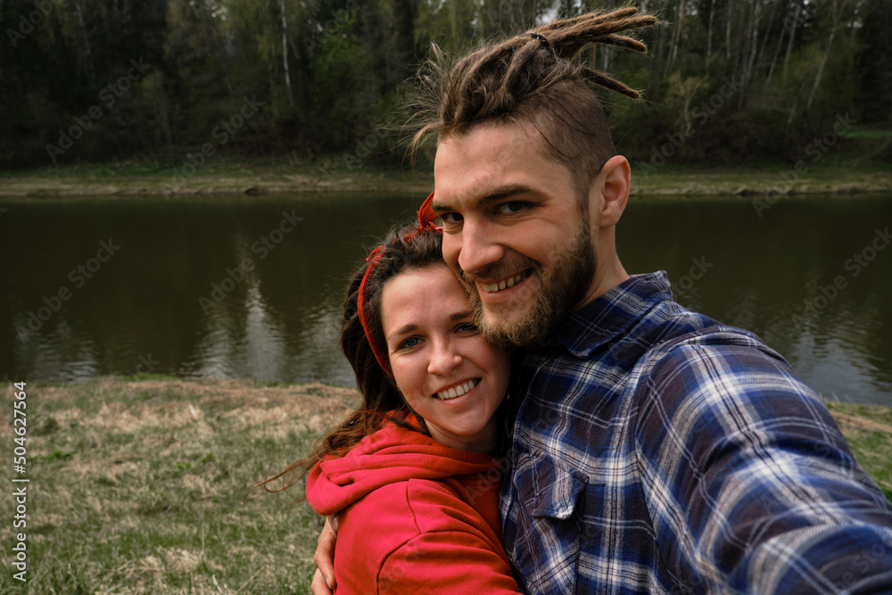 Concept of support and family values. Young European couple, man and woman with dreadlocks hug and take selfie in spring park. Cheerful people walk by river in summer and smile. Concept of support and family values. Young European couple, man and woman with dreadlocks hug and take selfie in spring park. Cheerful people walk by river in summer and smile.