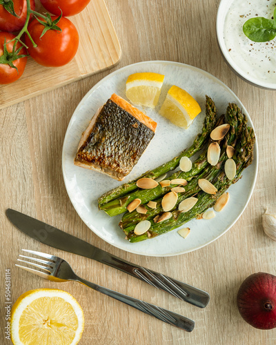 Salmon with grilled asparagus sprinkled with roasted almond flakes, surrounded by tomatoes, lemon, red onion and garlic sauce in a bowl. Flat view photography of foo.