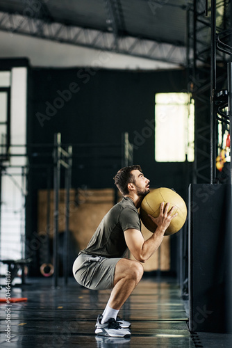 Athletic man exercising squats while holding medicine ball during cross training in gym.