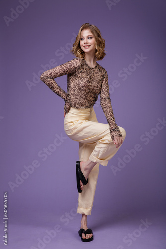 High fashion photo of a beautiful elegant young woman in a pretty beige cream linen trousers, pants, brown blouse with a spotted floral pattern on purple lilac background. Slim figure. Studio Shot