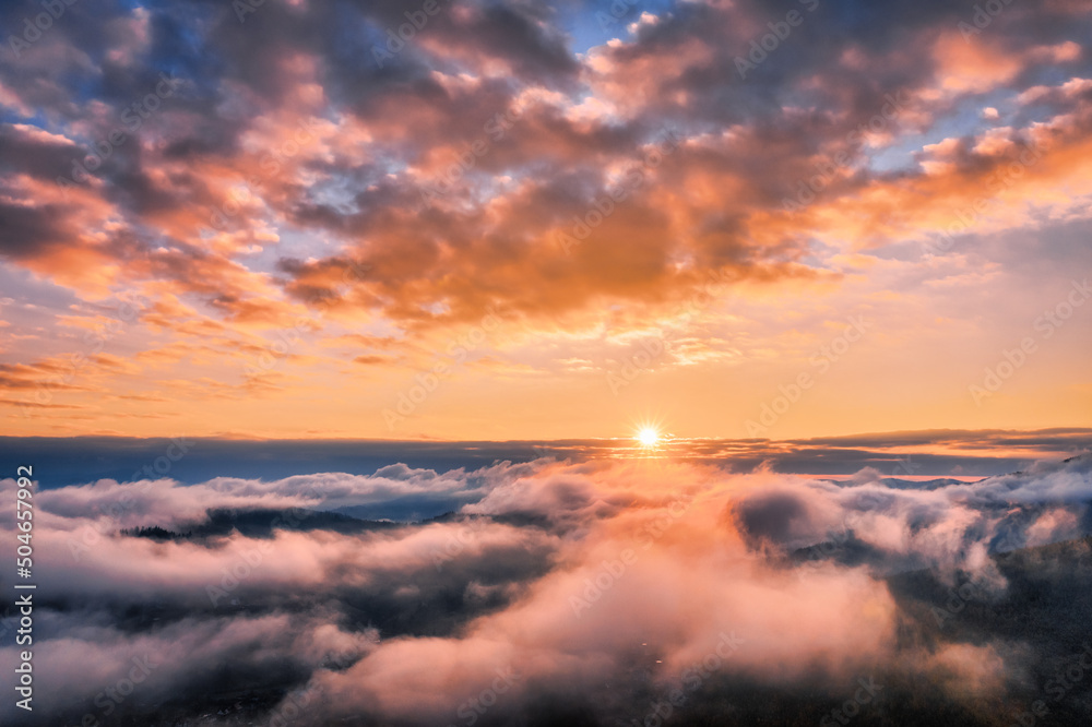 Mountains in low clouds at sunset in summer. Aerial view of mountain hills in fog. Beautiful landscape with high rocks, forest, colorful sky. View from above of mountain valley in clouds. Foggy hills