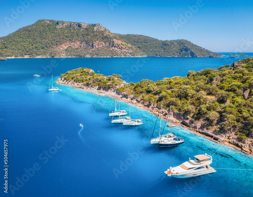 Aerial view of beautiful yachts and boats on the sea at sunset in summer. Gemiler Island in Turkey. Top view of luxury yachts, sailboats, clear blue water, beach, mountain and green forest. Travel