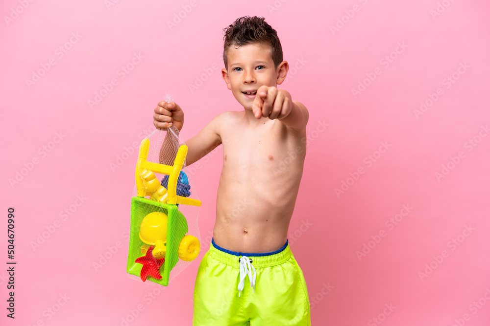 Little caucasian boy holding beach toys isolated on pink background points finger at you with a confident expression Little caucasian boy holding beach toys isolated on pink background points finger at you with a confident expression