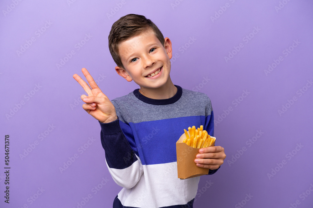 Little boy holding fried chips isolated on purple background smiling and showing victory sign Little boy holding fried chips isolated on purple background smiling and showing victory sign