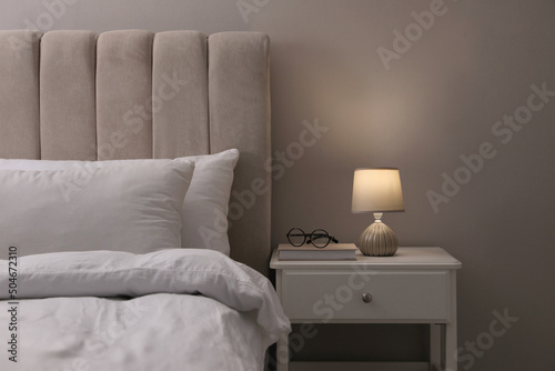 Stylish lamp, book and glasses on bedside table indoors. Bedroom interior elements