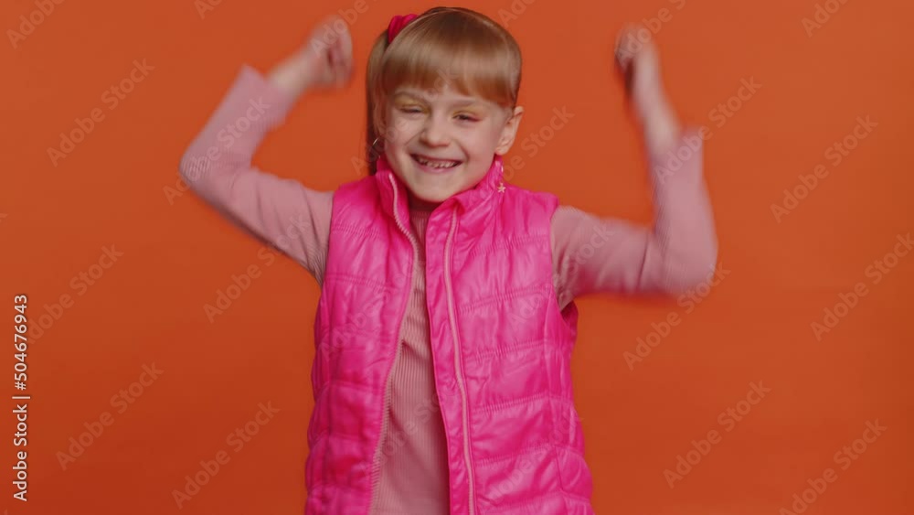Happy joyful girl in pink blouse shouting, raising fists in gesture I did it, celebrating success, winning, birthday, lottery goal achievemen. Little child kid indoors on orange studio wall background Happy joyful girl in pink blouse shouting, raising fists in gesture I did it, celebrating success, winning, birthday, lottery goal achievemen. Little child kid indoors on orange studio wall background