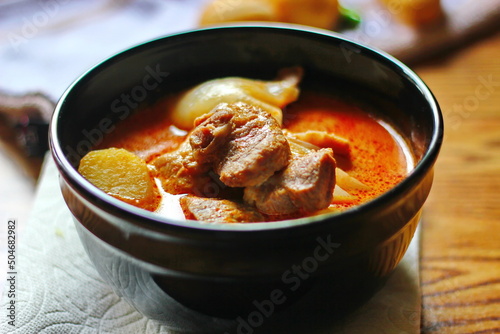 Massaman curry in black crockery served with boiled potatoes and chili on a brown wooden table.