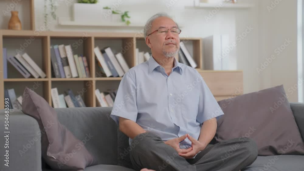 Asian senior man practicing yoga and meditating while sitting on the couch in lotus position and practicing breathing exercise at home. retirement senior male lifestyle living healthcare concept. Asian senior man practicing yoga and meditating while sitting on the couch in lotus position and practicing breathing exercise at home. retirement senior male lifestyle living healthcare concept.
