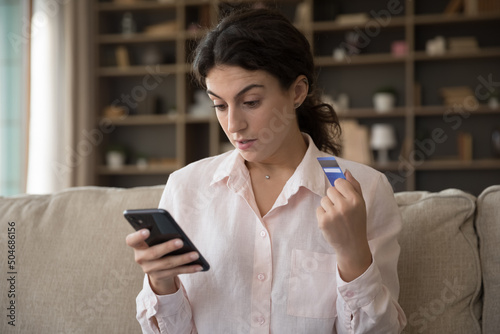 Young woman sit on sofa holds debit card staring at smartphone screen, feels surprised by insufficient funds, problem with online payment. Scam or unexpected discounts for goods on web store concept photo