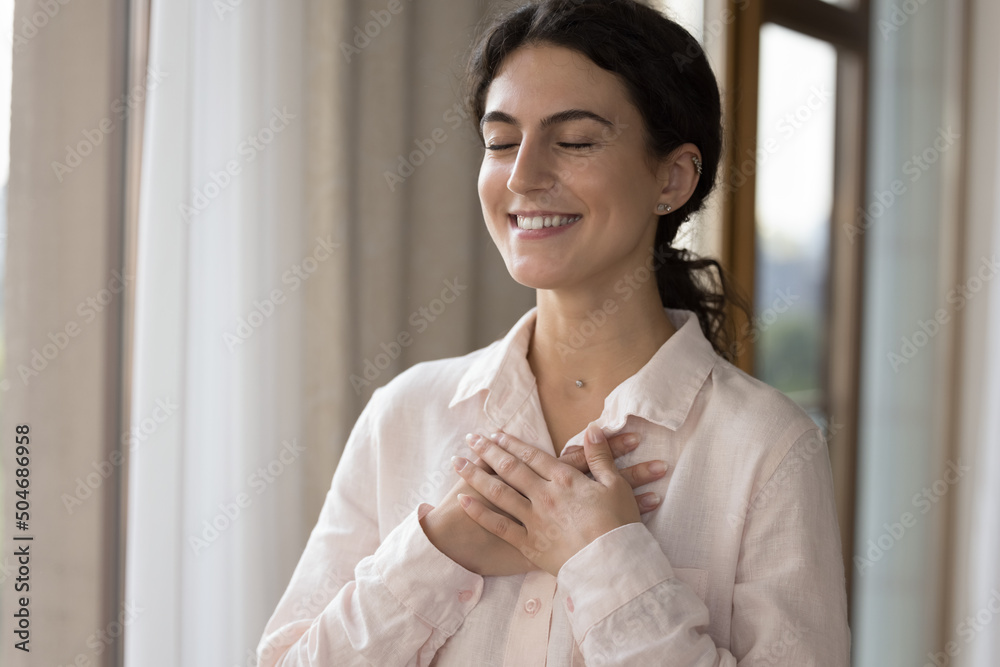 Young lovely woman standing alone indoors closed her eyes put folded palms on chest looks grateful, feeling deep appreciation, express sincere feelings. Believe, charity, symbol of gratitude concept Young lovely woman standing alone indoors closed her eyes put folded palms on chest looks grateful, feeling deep appreciation, express sincere feelings. Believe, charity, symbol of gratitude concept
