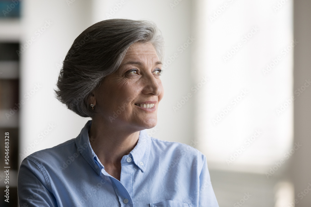 Happy pensive elder 60s woman looking out of window away with hope, thinking of good health, insurance benefits, family visit, dreaming of future, smiling at optimistic thoughts