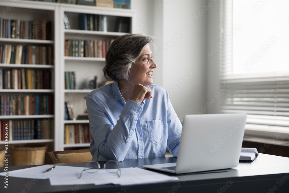 Happy pensive dreamy senior business lady enjoying job at workplace, thinking over future vision, project success, achievement, looking at window, smiling, sitting at table, laptop, papers