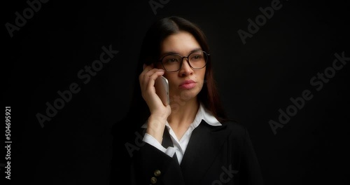 Business woman with glasses and long brown hair stands on an isolated black background in office clothes, answers a phone call, talks on a smartphone, says no with her head