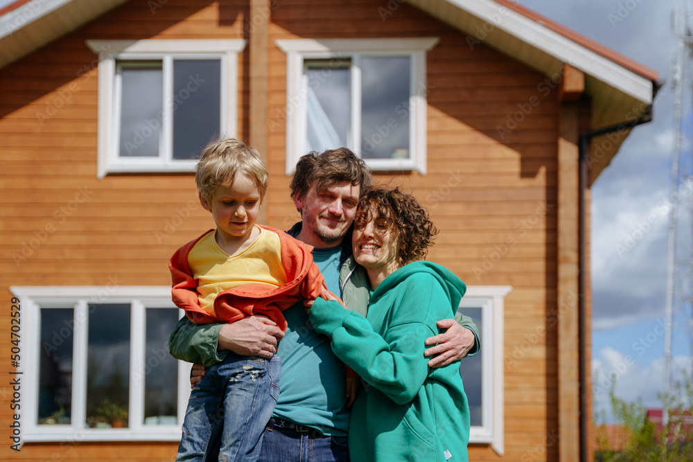 Happy family: Dad mom and their son hug, stand in front of their house, smile and look at the camera Happy family: Dad mom and their son hug, stand in front of their house, smile and look at the camera