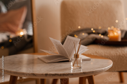 Liquid home fragrance in diffuser with open paper book on coffee table in bedroom indoors close up over scented candle. Cozy and hygge atmosphere. Aromatherapy. photo