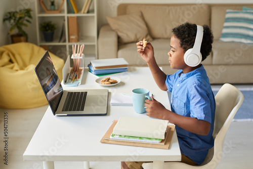 Side view of African-American boy in wireless headphones sitting at home desk and eating cookies while talking to online tutor © Mediaphotos