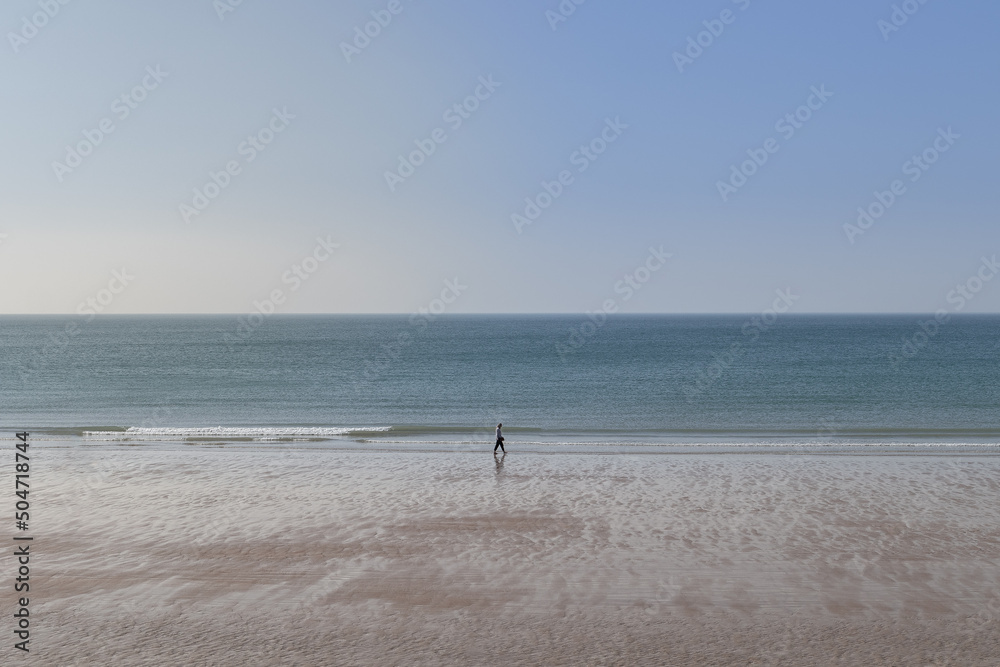 Promeneur sur la grande plage de St-Hélier sur l'île anglo normande de Jersey. La mer est calme et le ciel d'un grand bleu